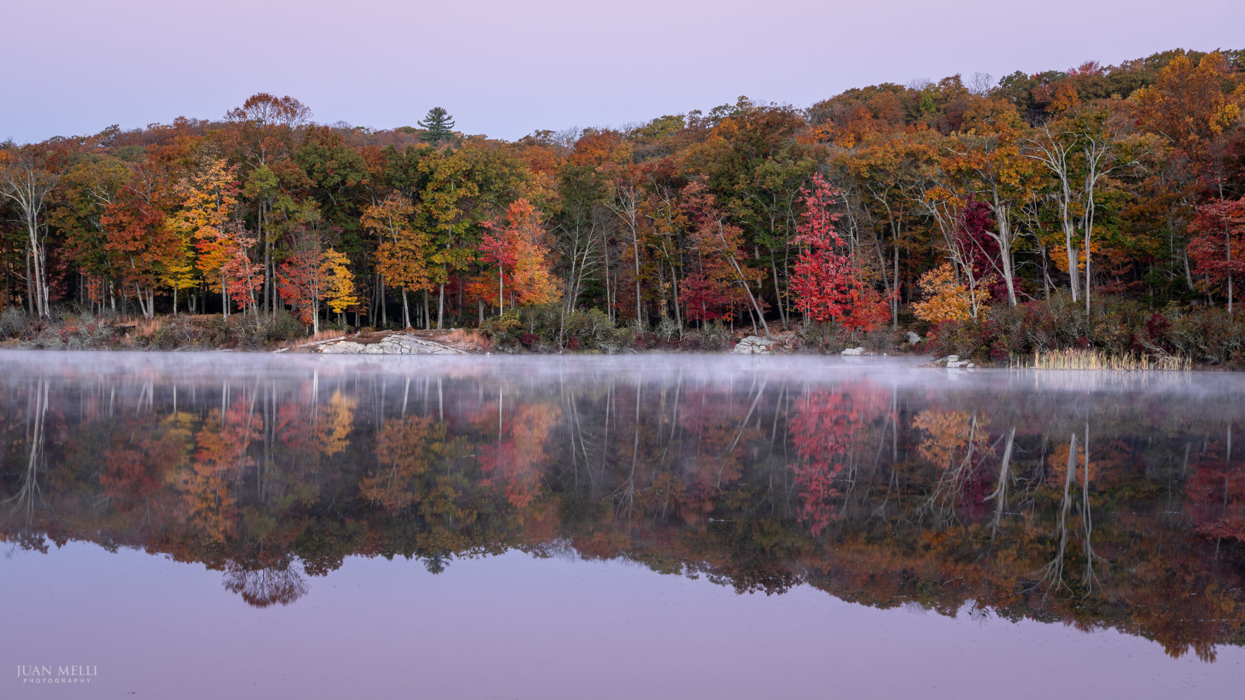 Fall foliage reflected in Lake Skannatati under a lavender dawn sky, with thin mist on the water