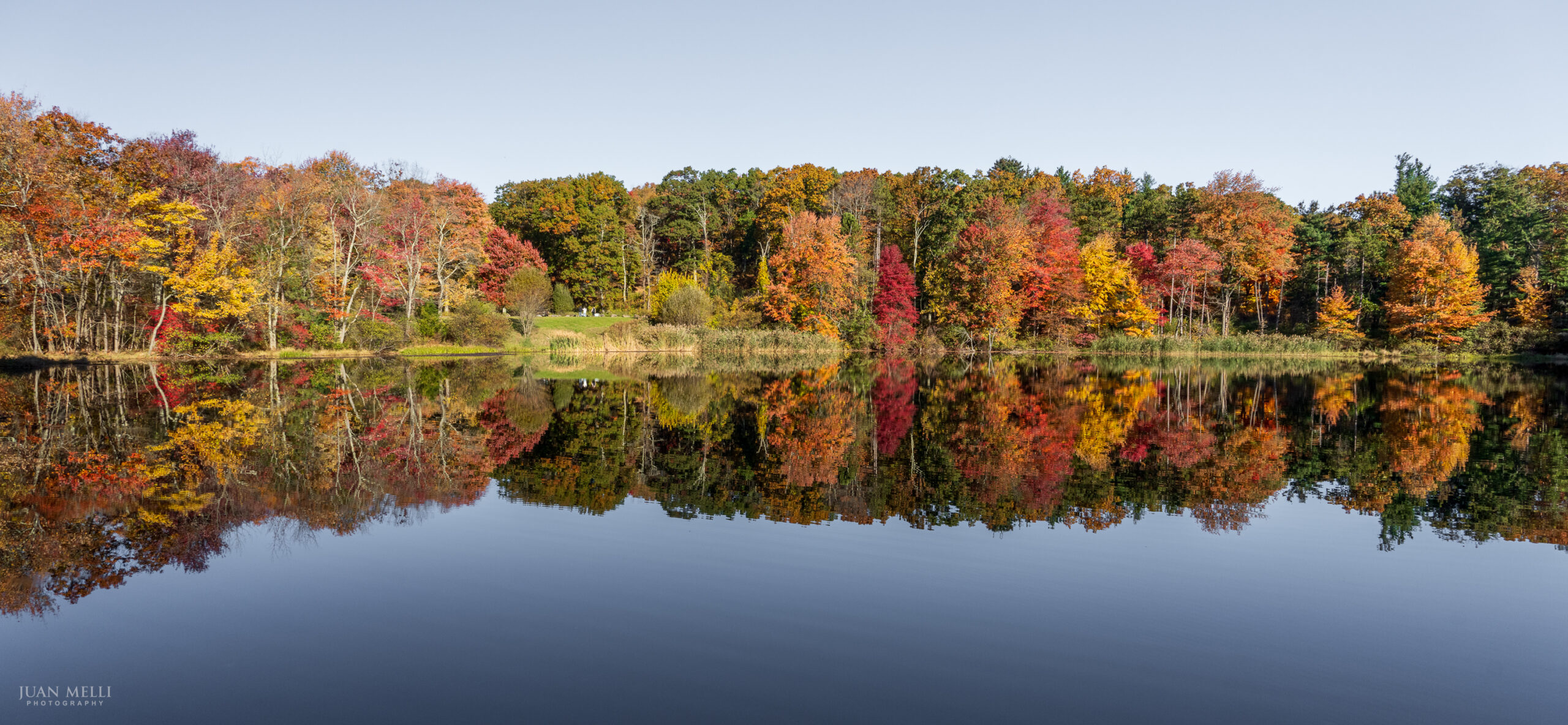 Peak fall color reflected in the still water of Cheesecote Pond under a clear blue sky