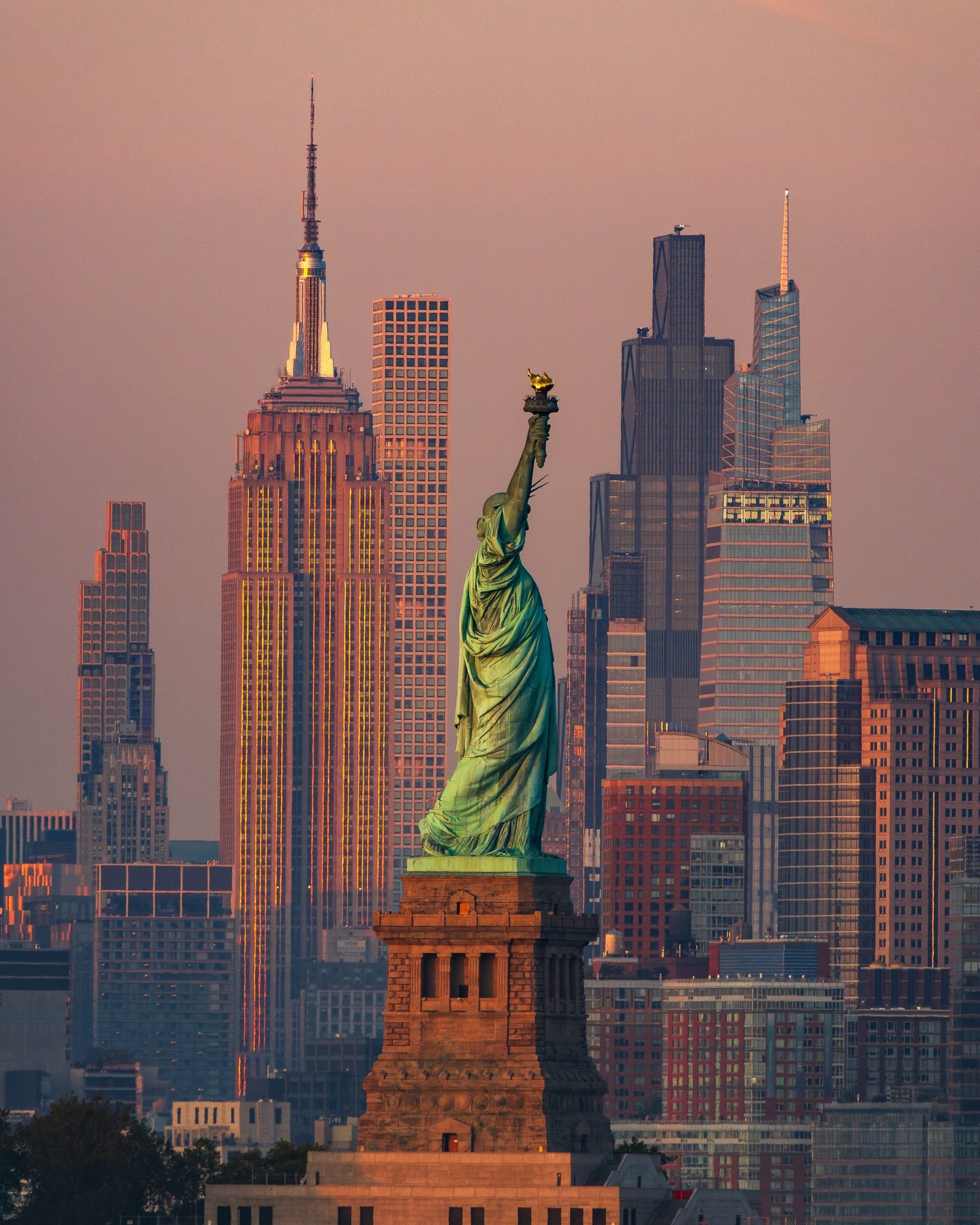 Photograph of the Statue of Liberty aligned with the Empire State Building and Midtown Manhattan at sunset.
