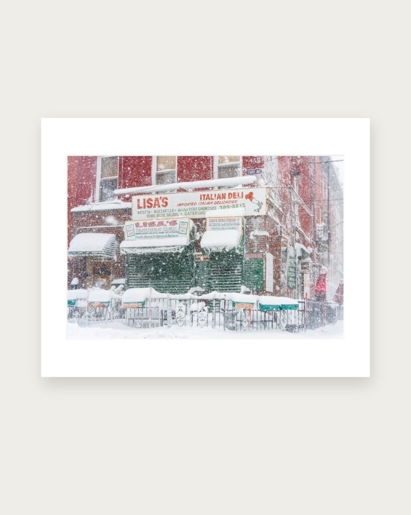Matted photographic print of Lisa’s Italian Deli in Hoboken during the Blizzard of 2026, its shutters closed and its signs and awnings heavy with snow.