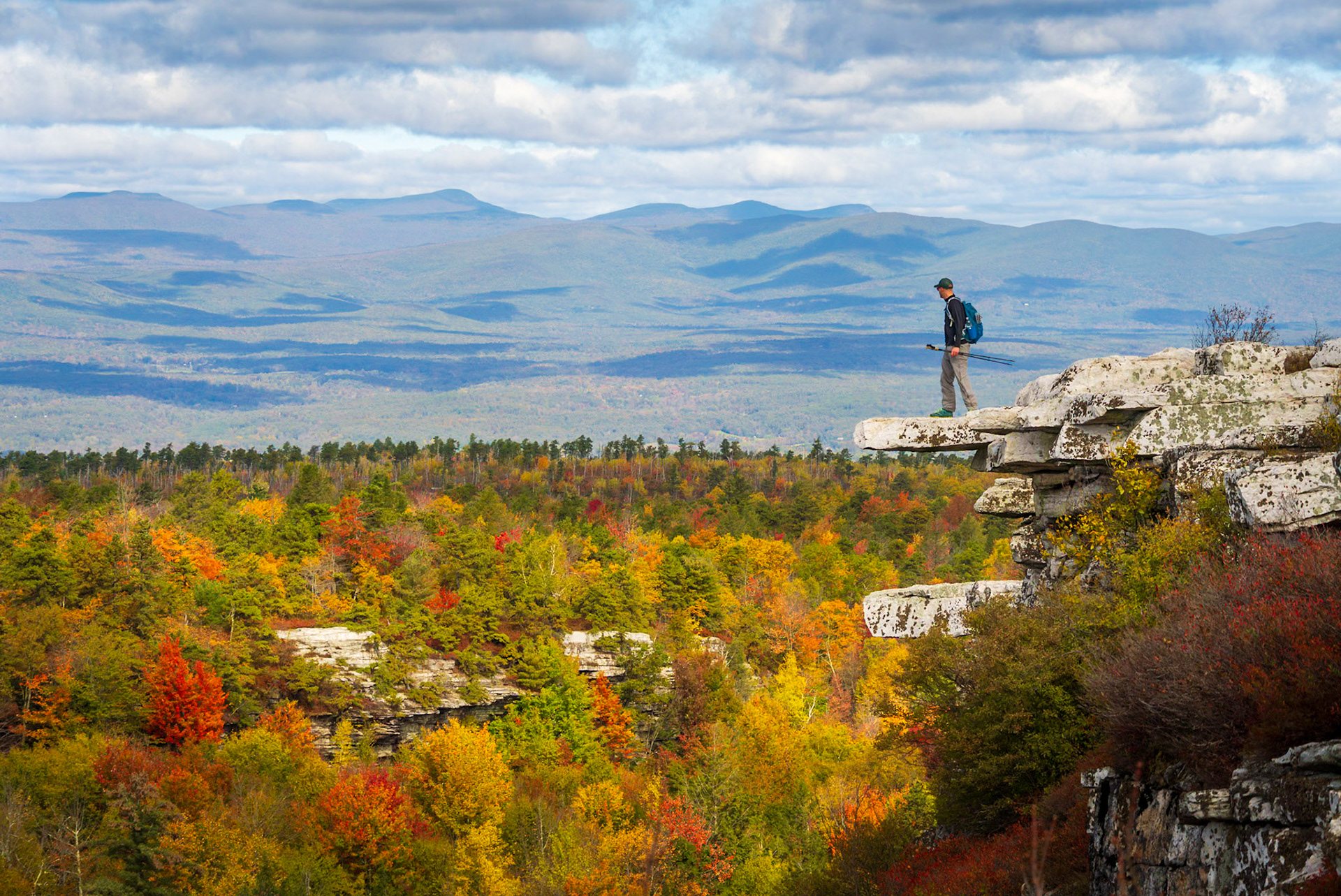 A hiker on a rock ledge on the Shawangunk Ridge above fall foliage
