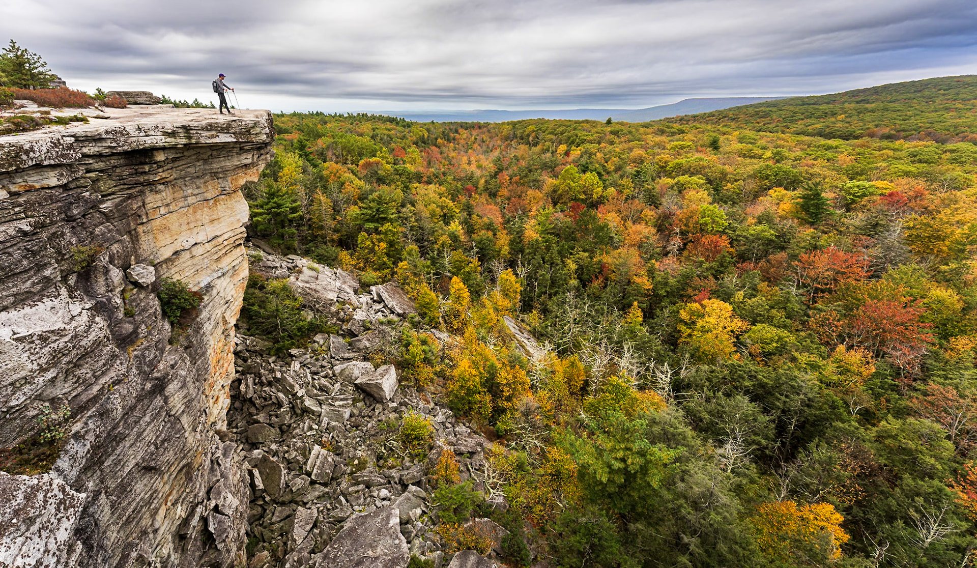 A hiker standing at the edge of a sheer cliff above autumn forest