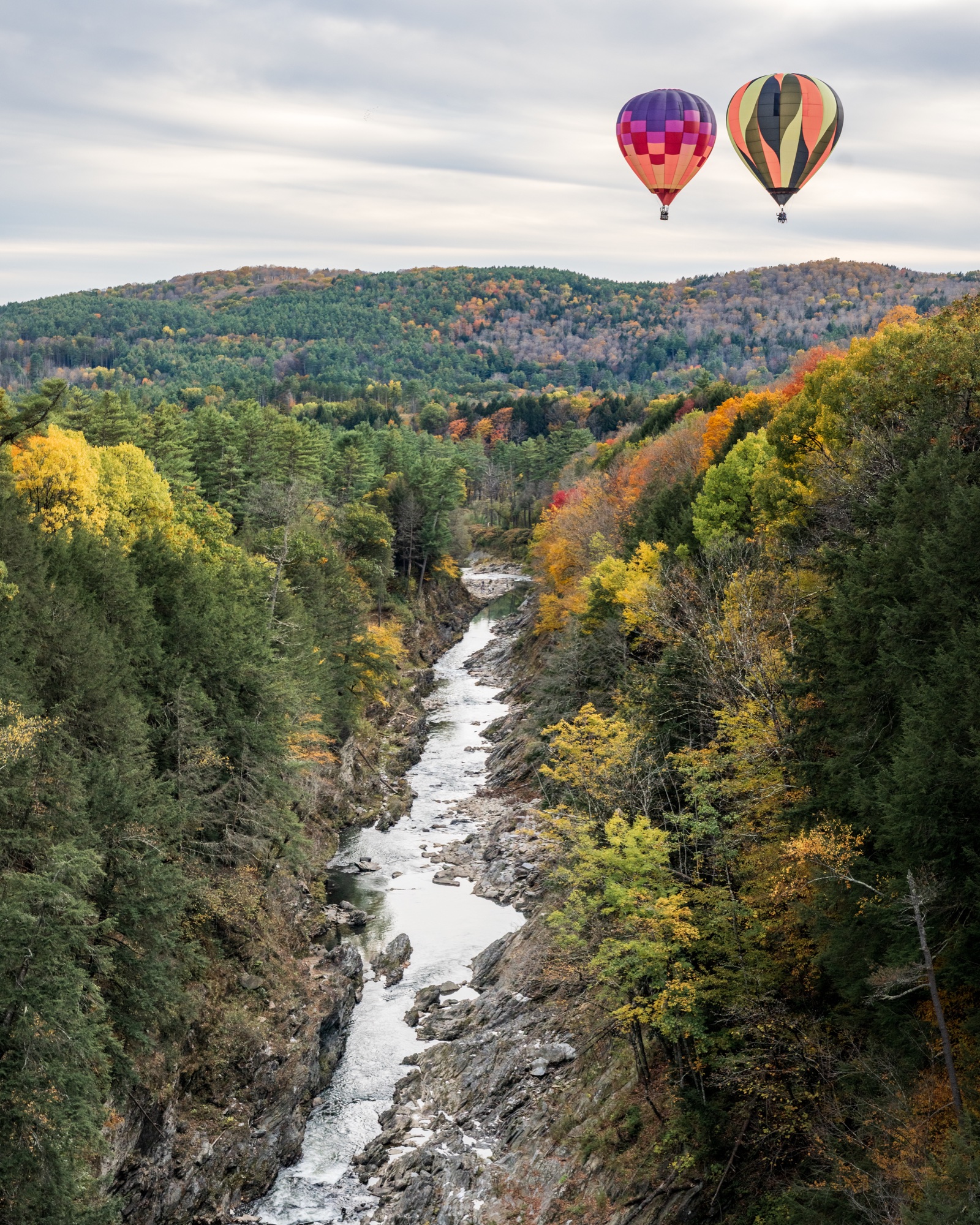 Two hot-air balloons above Quechee Gorge, with the Ottauquechee River far below and fall color on both rims