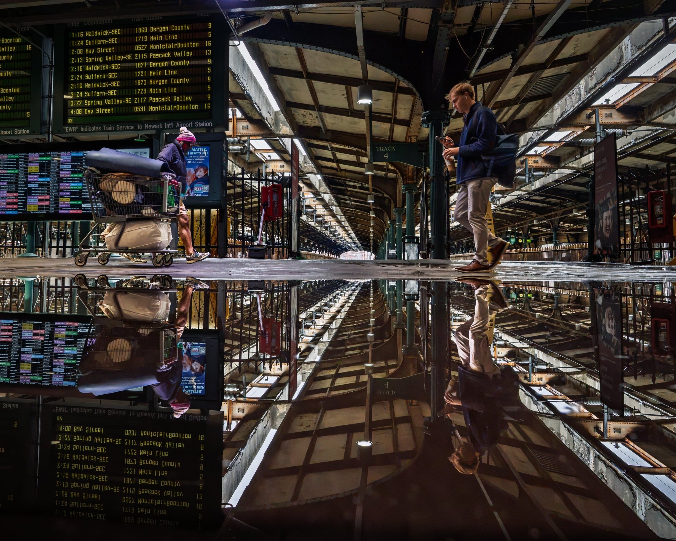 Photograph of Hoboken Terminal’s train shed reflected upside down in a rain puddle, with two travelers walking in opposite directions.