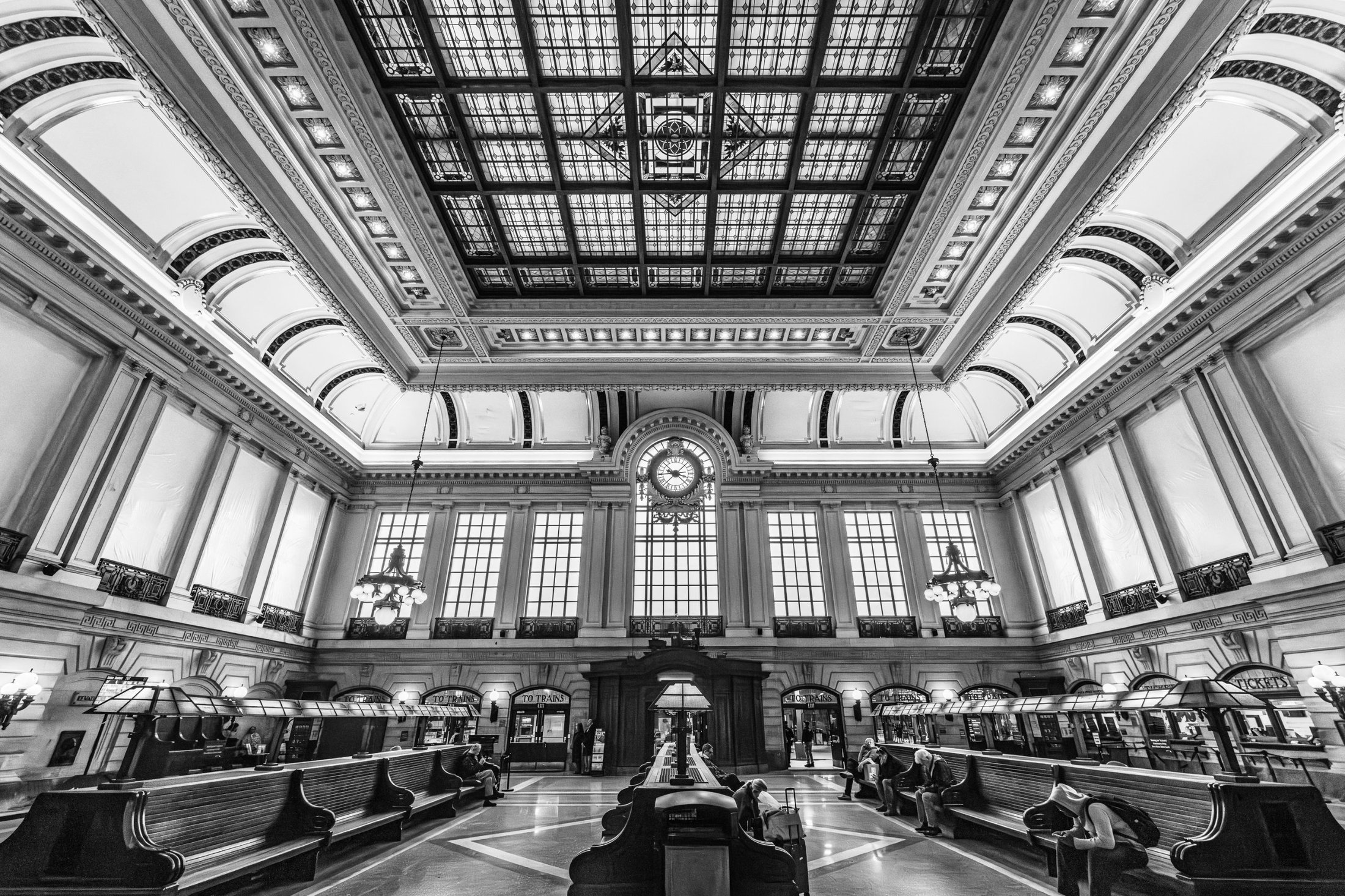 Black-and-white photograph of the grand waiting room at Hoboken Terminal, with its stained-glass ceiling, arched windows and long wooden benches.